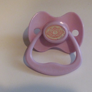 May include: A light pink baby pacifier with a rounded shield and a loop handle. The center of the pacifier features a circular design with a smiling sun graphic. The handle has the text "FOR BABIES".