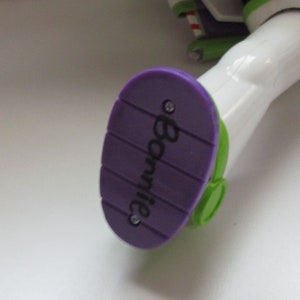 Puede incluir: Primer plano del pie de una figura de acci&oacute;n de Buzz Lightyear. El pie es morado con el nombre "Bonnie" impreso. La suela tiene un acento verde y la pierna de la figura es blanca.