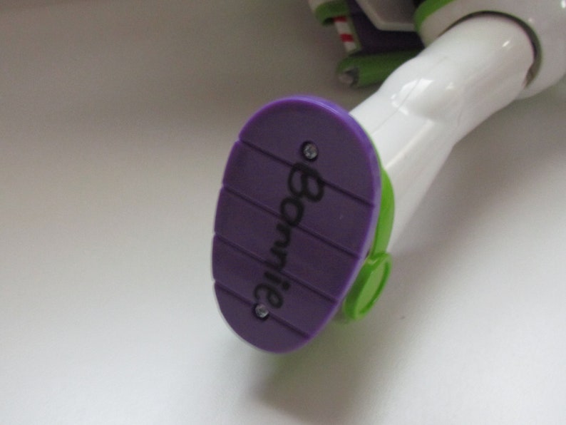 Puede incluir: Primer plano del pie de una figura de acci&oacute;n de Buzz Lightyear. El pie es morado con el nombre "Bonnie" impreso. La suela tiene un acento verde y la pierna de la figura es blanca.