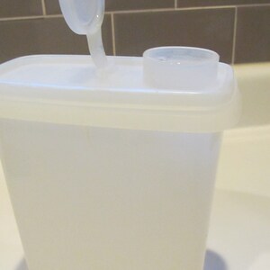 May include: A white plastic container with a flip-top lid and a small measuring cup on top. The container is rectangular with a slightly tapered design. The lid is open, revealing the interior.