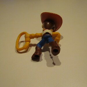 May include: A plastic toy figure of Woody from Toy Story. Woody is wearing his signature brown cowboy hat, blue jeans, and a yellow vest. He is holding a yellow lasso in his right hand.