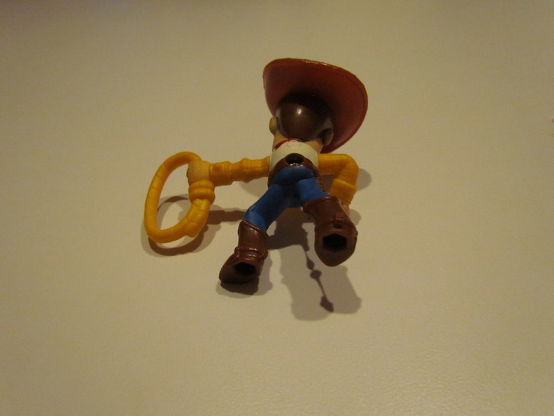 May include: A plastic toy figure of Woody from Toy Story. Woody is wearing his signature brown cowboy hat, blue jeans, and a yellow vest. He is holding a yellow lasso in his right hand.