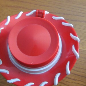 May include: A red plastic toy cowboy hat with a white wavy design around the brim. The hat has a hinged lid.