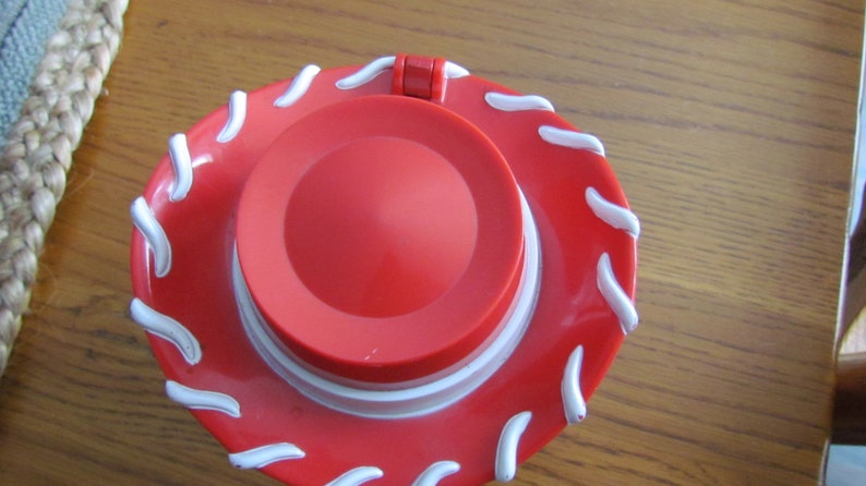 May include: A red plastic toy cowboy hat with a white wavy design around the brim. The hat has a hinged lid.