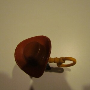 May include: A brown cowboy hat with a brown brim and a yellow plastic pull string. The hat is on a white surface.