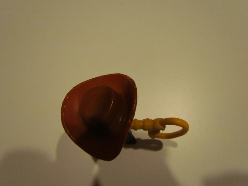 May include: A brown cowboy hat with a brown brim and a yellow plastic pull string. The hat is on a white surface.
