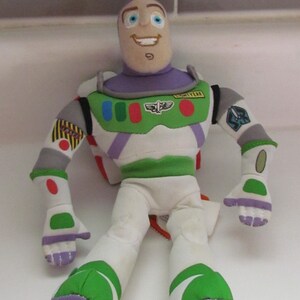 May include: A plush toy of Buzz Lightyear from the Toy Story franchise. The toy is wearing his signature space ranger suit with a purple chest piece, green and white legs, and a white helmet with a visor. The toy has a friendly face with blue eyes and a big smile. The toy is sitting on a white surface.