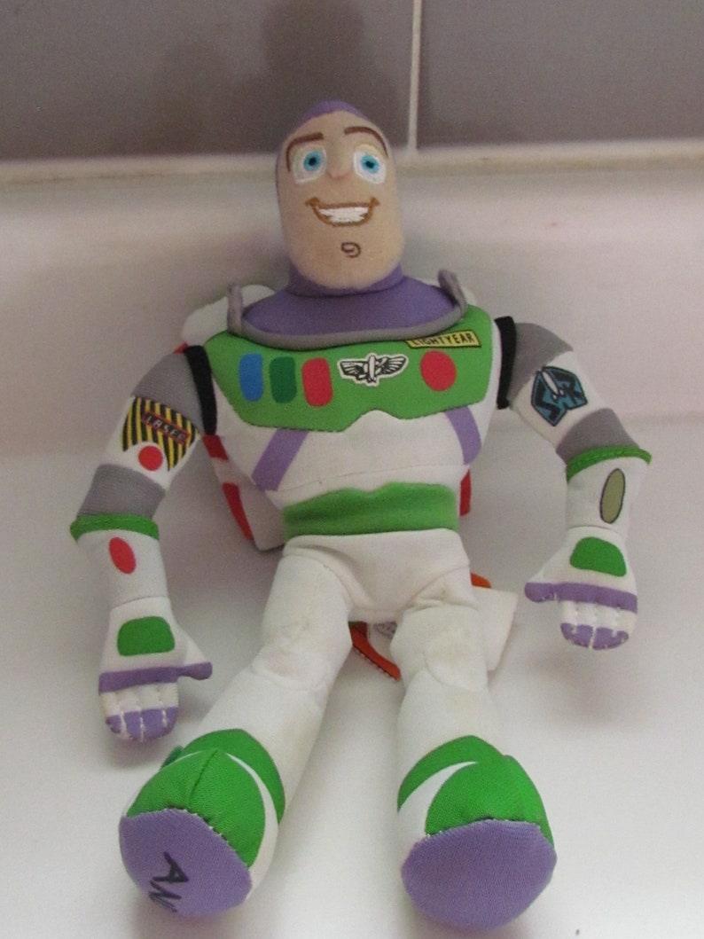 May include: A plush toy of Buzz Lightyear from the Toy Story franchise. The toy is wearing his signature space ranger suit with a purple chest piece, green and white legs, and a white helmet with a visor. The toy has a friendly face with blue eyes and a big smile. The toy is sitting on a white surface.