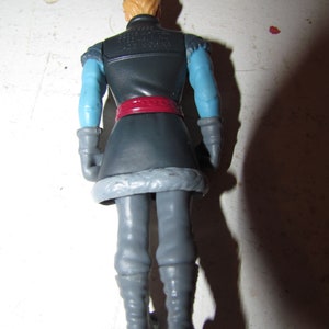 K&ouml;nnte beinhalten: Eine Kunststoff-Actionfigur einer Figur aus dem Film Frozen, von hinten gesehen. Die Figur hat blondes Haar, eine dunkelblaue M&uuml;tze und eine dunkelblaue Jacke mit hellblauen &Auml;rmeln. Ein roter G&uuml;rtel sowie graue Hose und Stiefel vervollst&auml;ndigen das Outfit.