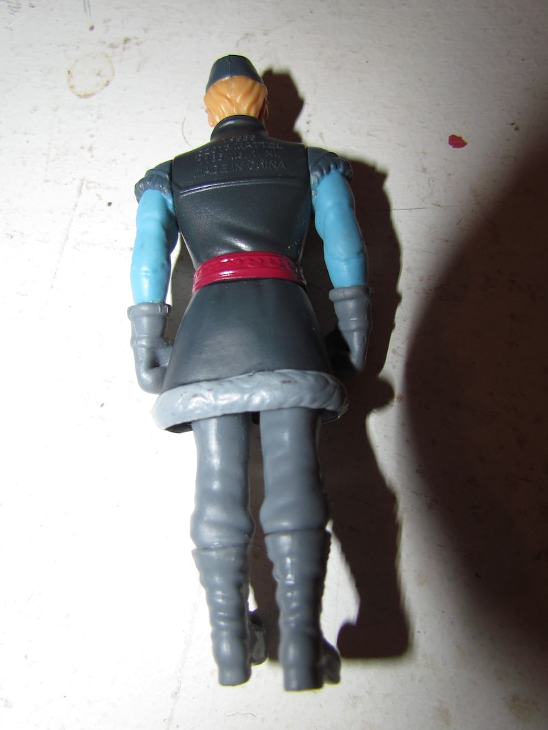 K&ouml;nnte beinhalten: Eine Kunststoff-Actionfigur einer Figur aus dem Film Frozen, von hinten gesehen. Die Figur hat blondes Haar, eine dunkelblaue M&uuml;tze und eine dunkelblaue Jacke mit hellblauen &Auml;rmeln. Ein roter G&uuml;rtel sowie graue Hose und Stiefel vervollst&auml;ndigen das Outfit.