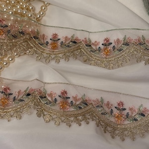 May include: Close-up of delicate embroidered lace trim with a scalloped edge. The trim features a floral design in pink, orange, and green, accented with gold thread. A strand of faux pearls rests on a white fabric background.