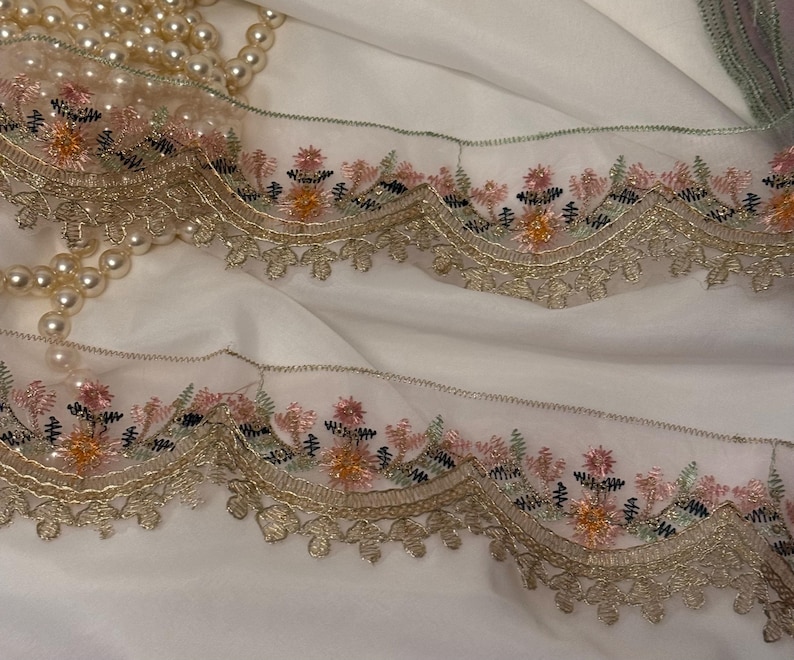 May include: Close-up of delicate embroidered lace trim with a scalloped edge. The trim features a floral design in pink, orange, and green, accented with gold thread. A strand of faux pearls rests on a white fabric background.