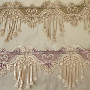 May include: Two sections of decorative lace trim. The top section features a beige and gold design with dangling tassels. The bottom section has a similar design, but with a mauve and beige color scheme. The lace has an ornate, detailed pattern.