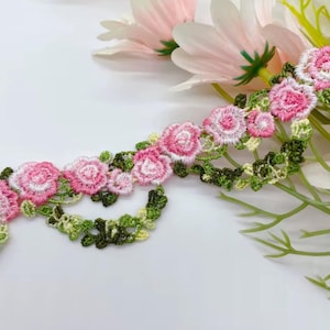 May include: Embroidered floral trim featuring pink and white roses with green leaves. The delicate trim is arranged on a white surface, with soft pink flowers in the background. This trim is suitable for various craft projects.