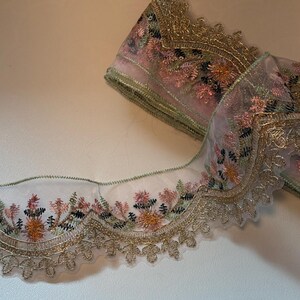 May include: A roll of decorative lace with a floral design. The lace features a sheer, light pink fabric with embroidered flowers in shades of pink, orange, and green. The edges are trimmed with gold metallic thread.