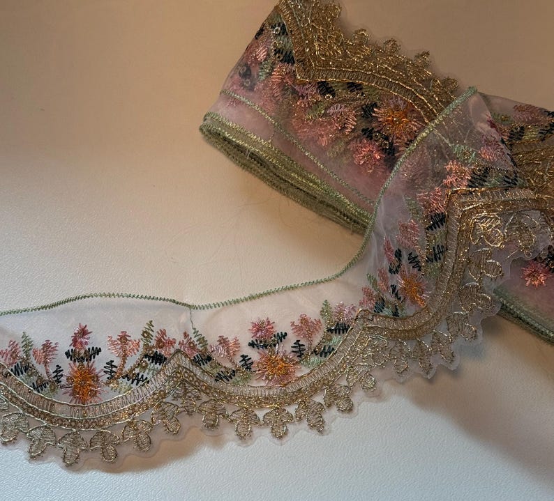May include: A roll of decorative lace with a floral design. The lace features a sheer, light pink fabric with embroidered flowers in shades of pink, orange, and green. The edges are trimmed with gold metallic thread.