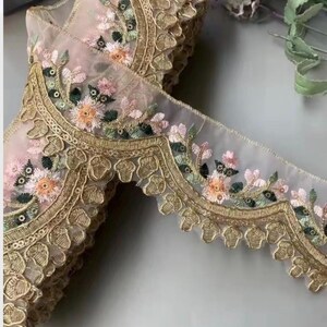 May include: Delicate floral embroidered lace trim in shades of pink, peach, green, and gold. The trim features a scalloped edge and intricate floral patterns, perfect for embellishing garments or crafts.