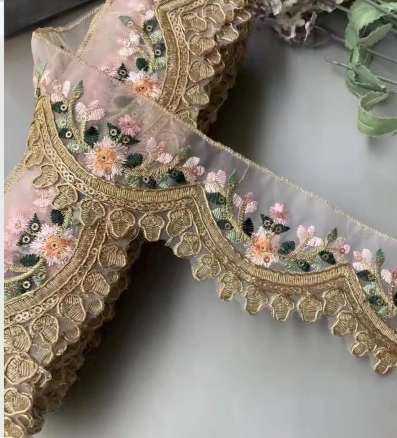 May include: Delicate floral embroidered lace trim in shades of pink, peach, green, and gold. The trim features a scalloped edge and intricate floral patterns, perfect for embellishing garments or crafts.