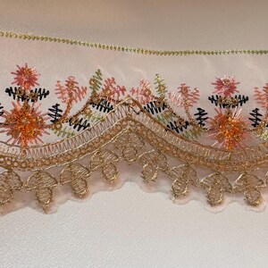 May include: Close-up of decorative lace trim featuring intricate floral embroidery. The design incorporates shades of gold, peach, pink, green, and black. The lace has a scalloped edge and is likely used for embellishing garments or crafts.