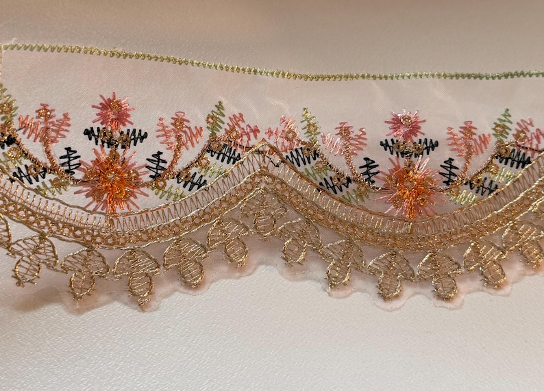 May include: Close-up of decorative lace trim featuring intricate floral embroidery. The design incorporates shades of gold, peach, pink, green, and black. The lace has a scalloped edge and is likely used for embellishing garments or crafts.