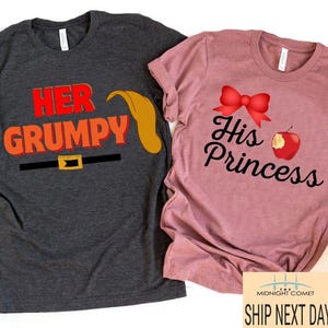 Op de afbeelding: Twee T-shirts, een grijs en een roze, met een bijpassend ontwerp voor koppels. Het grijze shirt zegt "Her Grumpy" met een cartoonafbeelding van een dwergenmuts en -baard. Het roze shirt zegt "His Princess" met een rode strik en een rode appel.