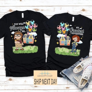 May include: Two black t-shirts with cartoon graphics. One shirt features a character with the text "You Will Always Be..." and a house with balloons. The other shirt says "My Greatest Adventure" with a character and house.