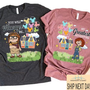 May include: Two t-shirts, one charcoal and one mauve, featuring cartoon graphics. The charcoal shirt says "You Will Always Be..." with a character, house, and balloons. The mauve shirt says "My Greatest Adventure" with a character, house, and balloons.