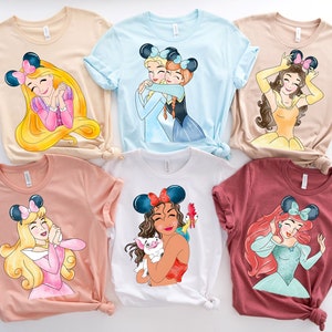 May include: Six different t-shirts featuring Disney princesses with mouse ears. Each shirt is a different color and features a different princess. The princesses are: Ariel, Belle, Cinderella, Mulan, Rapunzel, and Snow White.