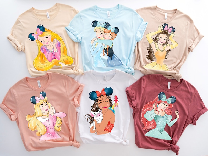 May include: Six different t-shirts featuring Disney princesses with mouse ears. Each shirt is a different color and features a different princess. The princesses are: Ariel, Belle, Cinderella, Mulan, Rapunzel, and Snow White.
