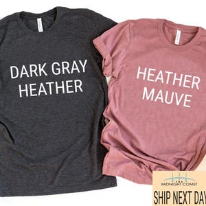 May include: Two t-shirts, one dark gray heather and one heather mauve, are displayed. Each shirt has a crew neck and the words "DARK GRAY HEATHER" and "HEATHER MAUVE" printed on the front in white. A small graphic and text are visible at the bottom.