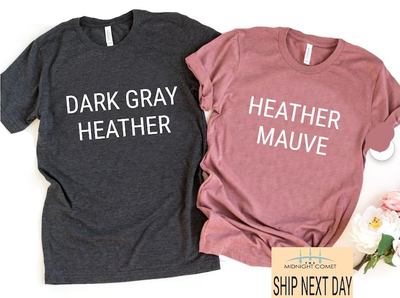 May include: Two t-shirts, one dark gray heather and one heather mauve, are displayed. Each shirt has a crew neck and the words "DARK GRAY HEATHER" and "HEATHER MAUVE" printed on the front in white. A small graphic and text are visible at the bottom.