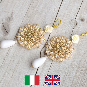 Può includere: Un paio di orecchini rotondi in perle dorate e bianche con un ciondolo a forma di fiore. Gli orecchini hanno un design delicato.