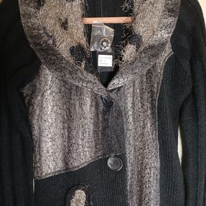May include: Black cardigan with a decorative silver and gray overlay. The sweater features a textured knit design, a shawl collar, and three buttons. A floral embellishment with a decorative button is on the lower left side.