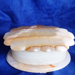 May include: A round, lidded box crafted from polished, translucent stone with a cream and amber-colored banded pattern. The lid features a scalloped edge design, while the base is a smooth, circular disc supporting the cylindrical container body.