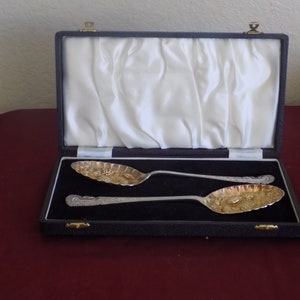 May include: Two silver serving spoons with gold accents in a black velvet lined box. The spoons have an ornate design and are in excellent condition.