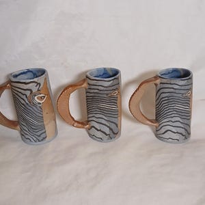 May include: Three handmade ceramic mugs with a unique design. Each mug features a cylindrical body with a textured, gray and white striped pattern, a brown handle, and a blue interior. The mugs are displayed on a white surface.