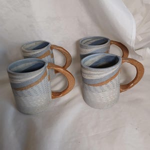 May include: Four ceramic mugs with a light blue and tan color scheme. The mugs have a textured surface with a tan handle and a blue and tan stripe around the middle. The mugs are handmade and have a rustic feel.