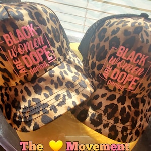 May include: Two brown and black leopard print trucker hats with black mesh back panels. The hats have pink text that reads "Black women are dope period."  The text is in a square box. The hats are sitting on a yellow surface. The text "The ❤️ Movement" is visible at the bottom of the image.