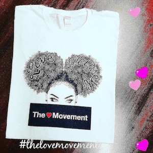 May include: White t-shirt with a black and white illustration of a woman with two afro puffs. The text "Love your self" is written on the puffs. The text "The ♥ Movement" is written on a black banner below the illustration. The hashtag #thelovemovement is written below the banner.