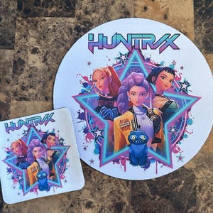 May include: A white round mousepad and a square coaster featuring a colorful graphic. The graphic includes the text "HUNTRAX" and illustrations of three women and a blue creature inside a star-shaped frame with a neon blue and pink color scheme.