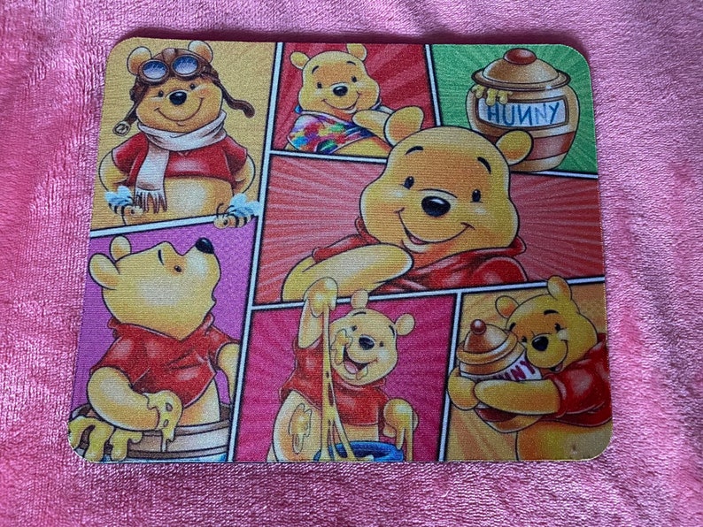 May include: A colorful mouse pad featuring Winnie the Pooh and his friends. The pad has a comic book style design with different scenes of Winnie the Pooh and his friends. The text "Hunny" is featured on the pad.