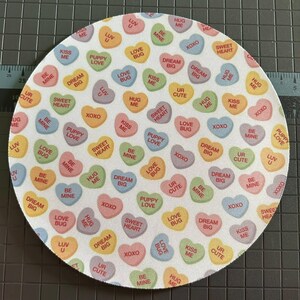 May include: A round, white surface covered in colorful candy hearts. Each heart has a different pastel color and a sweet message like "Kiss Me," "Love Bug," and "Sweet Heart." The overall design is playful and romantic.