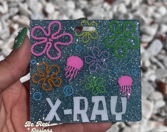Pode incluir: Um chaveiro de glitter azul turquesa com o texto "X-RAY" em branco e desenhos de água-vivas e flores de desenhos animados coloridos.
