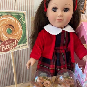 Puede incluir: Una muñeca con una chaqueta roja y un vestido a cuadros está de pie junto a una exhibición de mini bagels. La exhibición tiene cuatro cúpulas de plástico transparente con bordes dorados, cada una con varios mini bagels. Un letrero sobre la exhibición dice "classic Bagel rolling your day!"