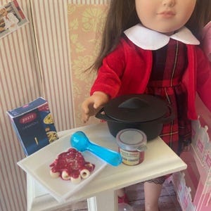 Könnte beinhalten: Eine Puppe in einem rot-schwarz karierten Kleid und einer roten Jacke sitzt an einem Tisch mit einem weißen Teller Pasta, einem blauen Löffel, einem kleinen Glas Pastasoße und einem schwarzen Topf. Die Schachtel Pasta trägt die Aufschrift "pasta" und "crespelle".