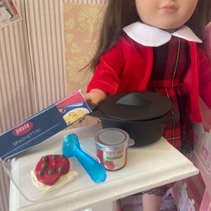 May include: A doll wearing a red and black plaid dress and a red jacket is setting the table with a pot, a jar of tomato sauce, a blue spoon, and a plate of spaghetti. The table is white and has a wooden base.