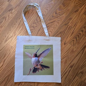 May include: A white canvas tote bag with a green background and a photo of a broad-tailed hummingbird in flight. The text "BROAD-TAILED HUMMINGBIRD" is printed in black at the top of the photo.