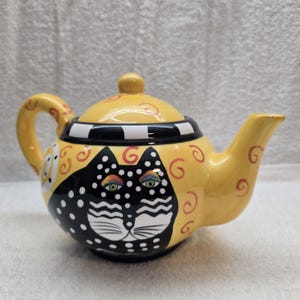 May include: A whimsical yellow teapot with a black cat face design. The teapot features a black and white striped band around the middle, a yellow lid, and a curved handle. The cat's face has green and rainbow-colored eyes.