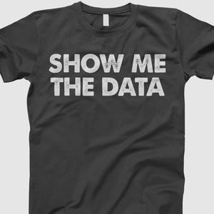 May include: A dark gray t-shirt with the phrase "SHOW ME THE DATA" printed in distressed white lettering. The shirt has a crew neck and short sleeves, suitable for casual wear.