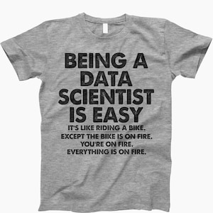 May include: Gray t-shirt with a black graphic that reads "Being a Data Scientist is Easy. It's like riding a bike. Except the bike is on fire. You're on fire. Everything is on fire."